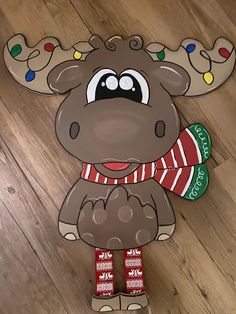 a paper moose with christmas lights on it's antlers and his scarf is standing in front of a wooden floor