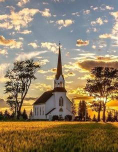 a church in the middle of a field with trees and clouds above it at sunset