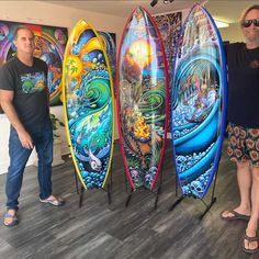 two men standing next to three surfboards in front of paintings on the wall behind them