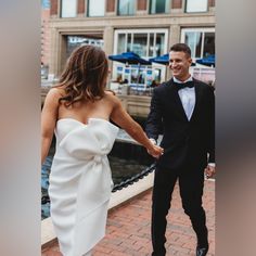 a bride and groom hold hands as they walk down the street in their tuxedos