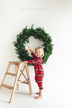 a little boy standing in front of a christmas wreath on a step ladder and pointing at it