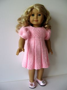 a doll with blonde hair wearing a pink knitted dress and white tennis shoes, standing against a wall