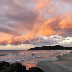 people are walking on the beach as the sun goes down over the water and clouds