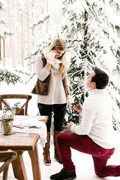 a man kneeling down next to a woman on top of a snow covered ground in front of a tree