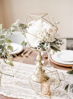 the table is set with white flowers and greenery in geometric vases on top of each other