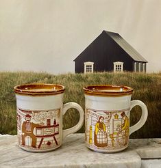 two coffee mugs sitting next to each other on a stone slab with a house in the background