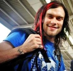 a man with long red hair is smiling at the camera while holding onto his backpack