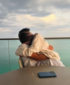 a man is hugging his friend while sitting at a table with the ocean in the background