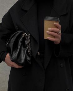 a woman in a black coat holding a coffee cup and a handbag on her hip