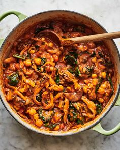 a pot filled with pasta and spinach on top of a marble countertop next to a wooden spoon