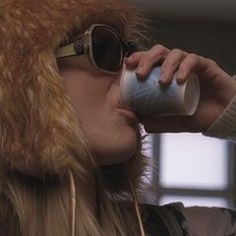 a woman drinking out of a cup while wearing a fur hat and sunglasses on top of her head