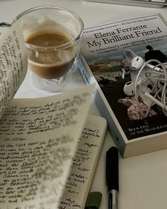 a cup of coffee next to an open book and a pen on a white table