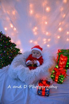 a baby in a santa hat is sitting next to presents and christmas lights on the wall