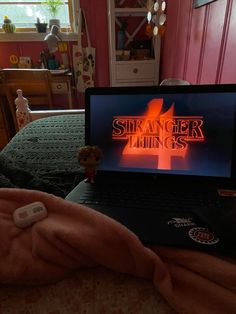 a laptop computer sitting on top of a bed next to a pink wall with the word strange things written on it