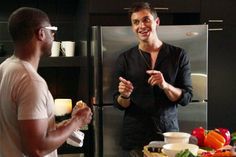 two men are standing in front of a refrigerator and talking to each other while eating food