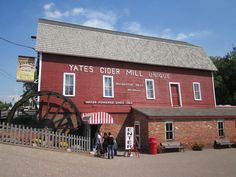 people standing in front of a red building with a water wheel on the outside and sign that says tates cider mill unique