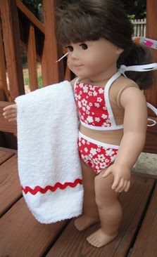 a doll is holding a towel on a wooden bench outside in the sun, wearing a red and white bathing suit