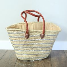 a woven basket with leather handles sitting on a wooden floor next to a white wall