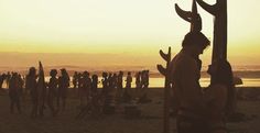 a group of people standing on top of a beach next to the ocean at sunset