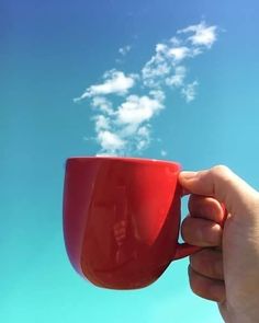 a hand holding a red coffee cup in front of a blue sky with white clouds