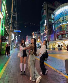 three girls are standing on the sidewalk in front of some buildings at night, and one girl is holding her hand up