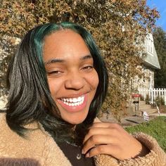 a woman with green hair smiling in front of a white picket fence and house behind her