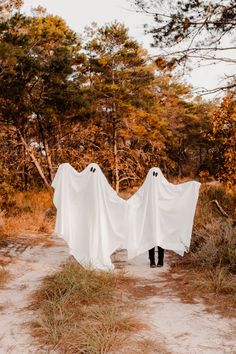 two people in white ghost costumes walking through the woods with their hands on each other's backs