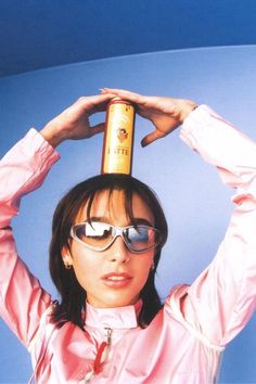 a woman with sunglasses and a pink shirt is holding a bottle on her head while standing in front of a blue background