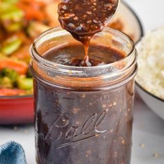 a spoon full of bbq sauce being poured into a jar with rice and vegetables in the background