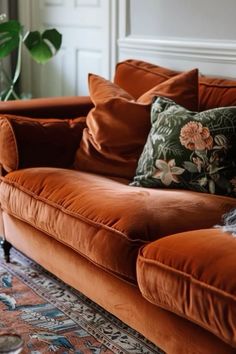 an orange couch with many pillows on top of it in front of a rug and potted plant