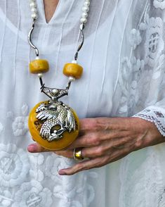 The majesty of the Himalayas. Tibetan yellow copal pendant embossed with Tibetan silver images of the Phoenix and the Elephant. Hill Tribe pure silver (.999) spacers, white fresh water pearls, vintage Tibetan copal beads, sterling silver beads, sterling silver clasp.  Length: 29" Pendant: 4"  Powerful and unique, this 29" long necklace centers on a large yellow Tibetan copal pendant, which is embossed with Tibetan "silver". A two-sided pendant, one side depicts the Elephant, which represents strength of mind in Tibetan Buddhism, the other side depicts the Phoenix, which symbolizes tranquility and peace. I have used vintage yellow Tibetan copal beads, large white pearls, and large spacers made of Hill Tribe pure silver (.999). It is definitely a powerful necklace that exudes the vast spirit Sterling Silver Amulet Necklace With Carved Details, Unique Ceremonial Jewelry With Large Pendant, Yellow Necklaces With Intricate Design As Gift, Yellow Necklaces With Intricate Design For Gift, Yellow Intricate Design Necklaces For Gift, Carved Amulet Jewelry For Collectors, Carved Amulet Pendant Jewelry, Carved Amber Amulet Jewelry, Ceremonial Carved Amulet Jewelry