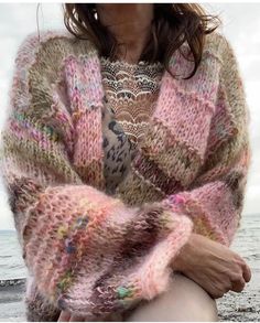 a woman sitting on top of a beach next to the ocean wearing a pink sweater