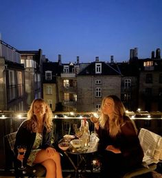 two women sitting at a table with wine glasses in front of them and lights on the balcony