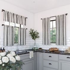 a kitchen with gray cabinets and black counter tops, white flowers on the table in front of two windows