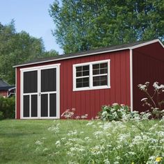 a red shed sitting in the middle of a lush green field with white flowers and trees