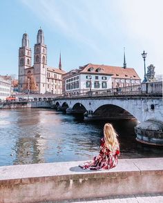 a woman sitting on the edge of a river looking at buildings and bridge over water