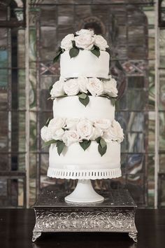 a three tiered cake with white flowers and greenery on the top is sitting on a table in front of a stained glass window