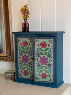 a blue cabinet with flowers painted on the front and sides, next to a mirror