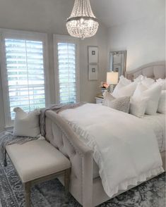 a white bed sitting in a bedroom next to a chandelier and window with shutters