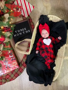 a baby wearing a red and black outfit laying next to a christmas tree with presents