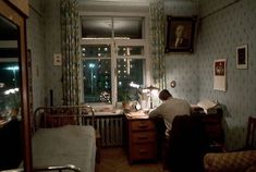 a man sitting at a desk in front of a window next to a bed and chair