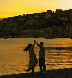 a man and woman holding hands while standing on the beach next to water at sunset