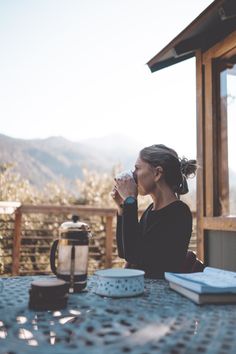 a woman sitting at a table drinking from a coffee cup with mountains in the background