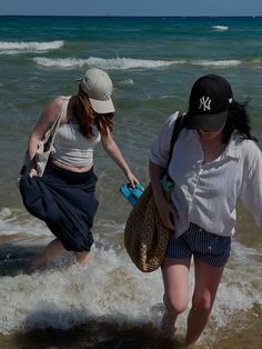 two women are walking into the ocean with their hats on and one is carrying a bag