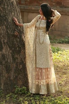 a woman standing next to a tree wearing a gold and beige dress with long sleeves