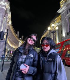 two women standing next to each other in front of a red double decker bus at night