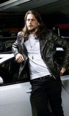 a man with long hair standing next to a car wearing a black leather jacket and white t - shirt