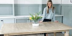 a woman standing next to a wooden table with flowers on it in a kitchen area