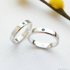 two white gold wedding bands with green cubics on each side, sitting next to a yellow flower