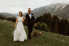 a bride and groom are walking on the grass in front of some mountains while holding hands